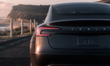 A sleek black Tesla Model 3 parked along a scenic coastline, with its logo and plate visible, under soft sunset lighting.