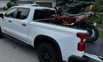 White Chevrolet Silverado pickup truck with black wheels transporting a red and black KTM motorcycle in its bed, parked in suburban driveway