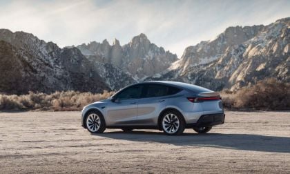 A silver 2026 Tesla Model Y Juniper parked on rocky terrain, with majestic snow-capped mountains in the background, under a clear blue sky.
