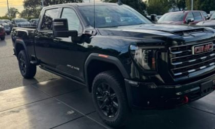 A sleek black GMC Sierra 2500 H truck is parked under an overcast sky, showcasing its bold grille and robust stance. Surrounding trees and cars set a serene backdrop.