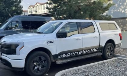 White Ford F-150 Lightning with "Aspen Electric" branding parked beside a van in a lot. Trees and residential buildings are visible in the background.