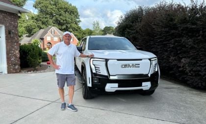 Perry is seen standing in a white polo shirt and cap smiling next to a white GMC Sierra EV in a suburban driveway. A brick house and greenery are visible in the background.