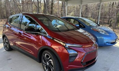 A maroon/red 2022 Chevrolet Bolt EUV is shown from a three-quarter front angle, parked next to a blue Nissan Leaf under a covered carport surrounded by trees.