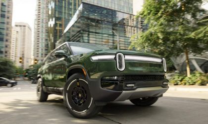 A sleek green electric SUV parked on a city street with modern skyscrapers in the background. Urban setting with greenery.