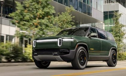 Dark green Rivian R1S SUV driving past modern glass building with blurred trees, front three-quarter view on urban street
