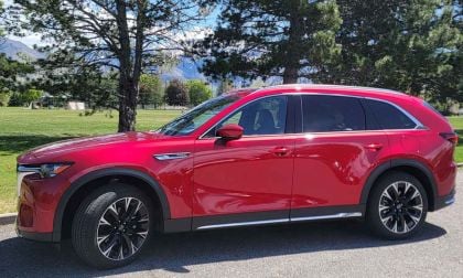 A Red 2025 Mazda CX-90 parked on a sunny day in a grassy area, with trees and mountains in the background.