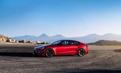 A red 2025 Tesla Model S parked on a winding road with mountains in the background under a clear blue sky.