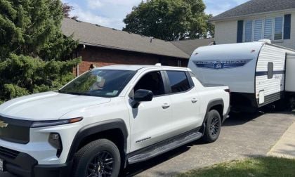 A white Chevrolet Silverado EV is parked in a driveway, hitched to a Gulf Stream travel trailer. A brick house and lush green trees are visible in the background.