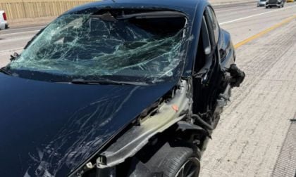 A black Tesla Model 3 on the side of the highway with a severely shattered windshield and damaged front, indicating a recent accident