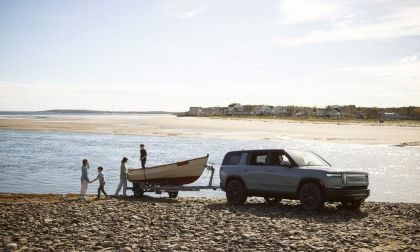 A family on a beach pulls a small boat from the water, with a Rivian R1S parked nearby under a clear sky.