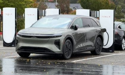 A Lucid Gravity GT is parked at a charging station on a rainy day. The futuristic vehicle is connected to a Tesla charger, surrounded by wet pavement and autumn trees.