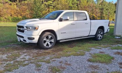 White 2024 Ram 1500 pickup truck shown from a front three-quarter view, parked on gravel with trees in the background, featuring chrome accents and alloy wheels.