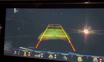 A car's rearview camera display shows a night scene with dark background and colorful guidelines for safe parking. A large lens flare is visible on the right.