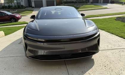 Sagar's 2024 grey Lucid Air Touring parked in a driveway, facing forward. The scene is suburban with neatly trimmed lawns and other cars in the background.