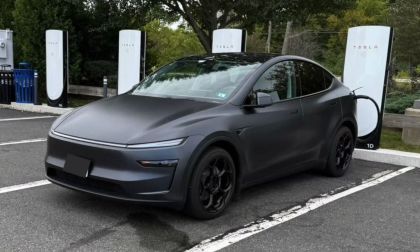 A matte black Tesla Model Y is parked at a Tesla charging station surrounded by greenery. The scene conveys a modern, eco-friendly vibe.