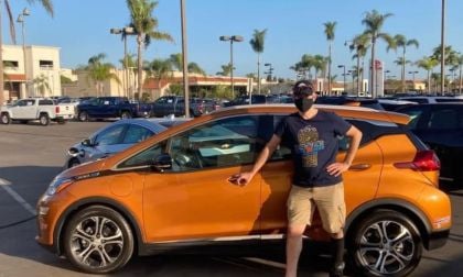 Scott wears a mask and is in casual clothing standing proudly beside an orange Chevy Bolt in a sunny parking lot filled with palm trees and parked vehicles.