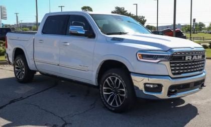 White Ram 1500 Limited parked outdoors, showcasing a shiny chrome grille and alloy wheels. The setting is a car lot with trees and blue sky in the background.