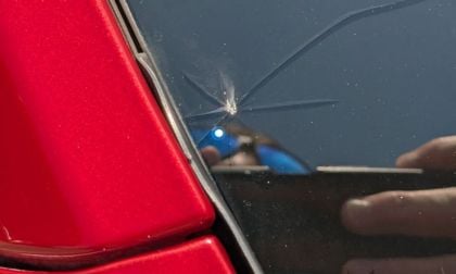 Close-up of Tesla Model Y's cracked windshield near a red frame with visible reflection of a person holding a phone. The tone suggests mild damage and concern.