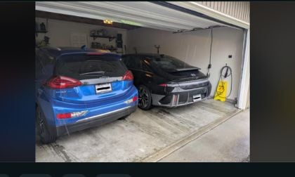 A blue Chevrolet Bolt EV and a black electric vehicle are parked in a garage, with cleaning supplies and shelves in the background.
