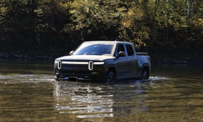 Rivian R1T electric pickup truck crossing a river, surrounded by autumn forest with water reflecting its metallic blue-gray body