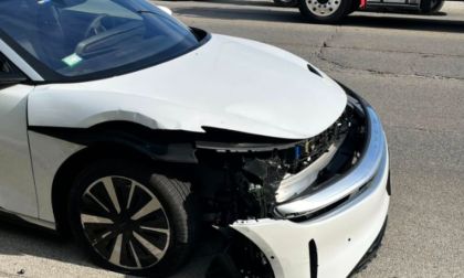 A white Lucid Air Touring with significant front-end damage, including a missing bumper and exposed internal components, is parked on a city street during daylight.