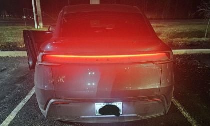 Rear view of a Tesla Model Y parked at night with the brake lights glowing red. The driver's door is slightly open, and the surroundings are dimly lit.