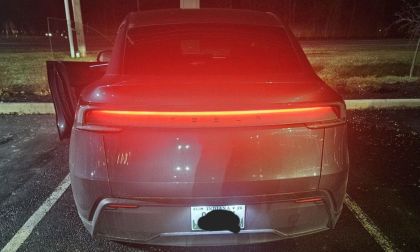 Rear view of a Tesla Model Y parked at night with the brake lights glowing red. The driver's door is slightly open, and the surroundings are dimly lit.