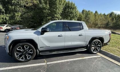 A grey GMC Sierra EV with large and stylish wheels is parked on a paved area. Surrounded by green trees, the scene conveys a modern, outdoorsy vibe.