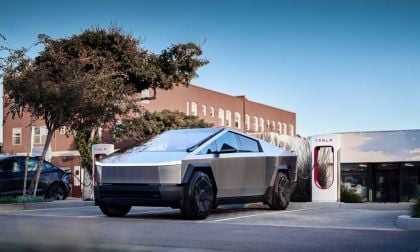 A Tesla Cybertruck parked at a charging station near a modern building, with trees and another Tesla vehicle in the background.