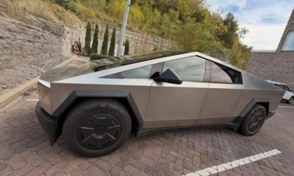 The futuristic and angular Tesla Cybertruck in a parking lot surrounded by a stone wall and greenery. The design is sleek and modern with a bold presence.