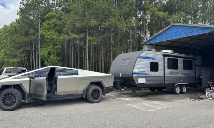 A 2025 Tesla Cybertruck parked next to a gray travel trailer under a metal shelter, surrounded by dense green trees.