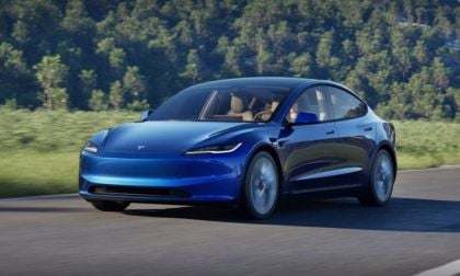 A blue Tesla Model 3 drives on a scenic road with lush greenery in the background, showcasing its sleek design and modern features.