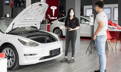 A Tesla sales representative explains features of a white Tesla Model 3 with the hood open, in a sleek showroom setting.
