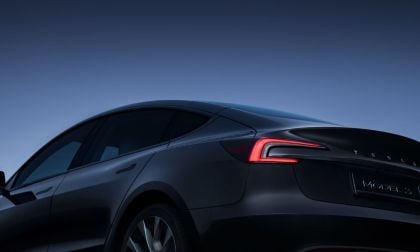 A dark gray Tesla Model 3 sedan shown from the rear three-quarter angle, highlighting its distinctive C-shaped LED taillights and sleek fastback profile against a deep blue background.