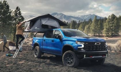 A blue Chevrolet Silverado 1500 truck with roof tent in a forested mountain area. A person climbs the tent ladder and another person assembles a ground tent nearby. Atmosphere is a peaceful outdoor scene.