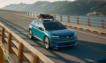 A turquoise 2026 Honda Prologue is shown from a three-quarter front angle driving across a bridge with mountains and a lake in the background, featuring a roof-mounted cargo carrier.