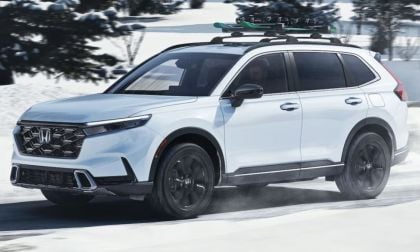 A white Honda CR-V drives on a snow-covered road with trees in the background. It has a roof rack carrying skis, conveying a sense of adventure and winter travel.