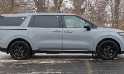 A grey Kia Carnival is shown from the side view, featuring black wheels and trim, photographed in a winter setting with snow-covered trees in the background.