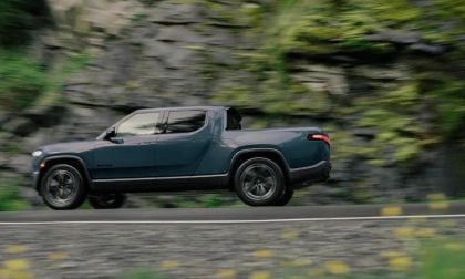 Dark blue electric pickup truck speeding along rocky mountain road with blurred green forest background