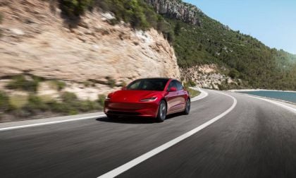 A red Tesla Model 3 speeds along a winding coastal road. Surrounded by rocky cliffs and greenery under a clear blue sky, the scene evokes freedom and adventure.