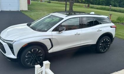 White Chevrolet Blazer EV SUV parked on black asphalt driveway with green lawn and trees in background, sleek modern design with black wheels