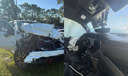 A totaled white GMC Sierra 1500 AT4 is shown in a grassy area, its front crumpled from a collision. The interior displays deployed airbags and a cracked windshield.