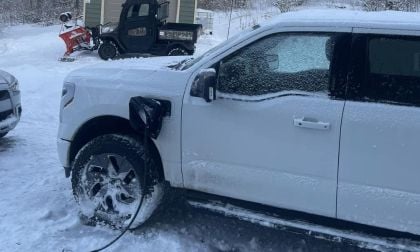 White Ford F-150 Lightning pickup truck shown from the side angle in snowy winter conditions, with snow-covered wheels and a utility vehicle with plow attachment visible in the background.