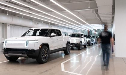 A row of white Rivian R1T in a modern workshop, with a blurred figure walking past under bright overhead lighting.