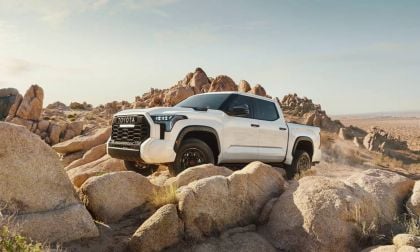 A white 2025 Toyota Tundra truck navigates rocky terrain under a clear sky, showcasing its rugged design and off-road capability.