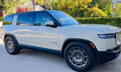 A Rivian electric SUV parked in a driveway, showcasing its sleek design, rugged tires, and modern details against a green backdrop.