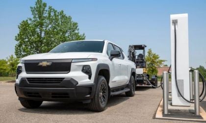 A white Chevrolet truck is parked beside an electric charging station, with green trees and a clear blue sky in the background.