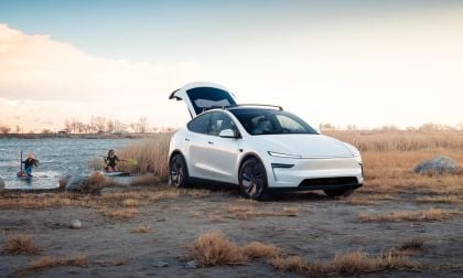 A white 2026 Tesla Model Y Juniper parked by a lake, with two people kayaking nearby under a colorful sky and tall grass.