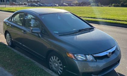 A dark silver 2009 Honda Civic is parked on a sunny street beside a green lawn. The car's exterior is shiny, and the setting is calm with a clear sky.