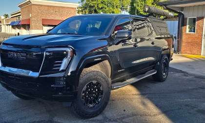 Black GMC Sierra Denali with off-road tires and an aftermarket bed cap parked outside a storefront.